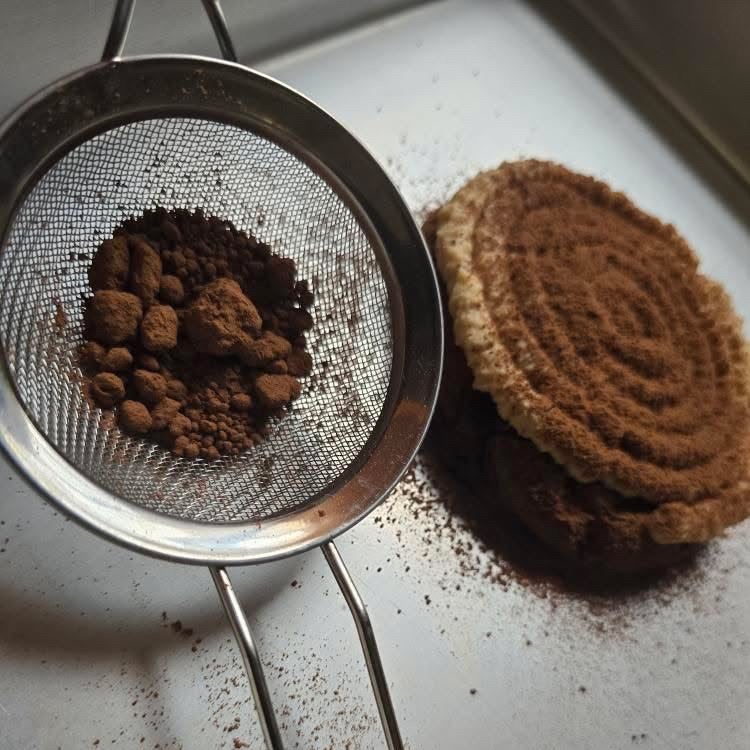 Metal mesh strainer with coffee grounds next to a brown coffee scrubber on a white surface.